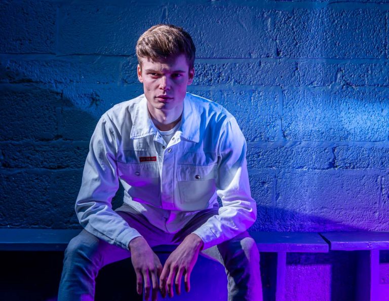 male model in white denim jacket sitting against a blue coloured brick wall